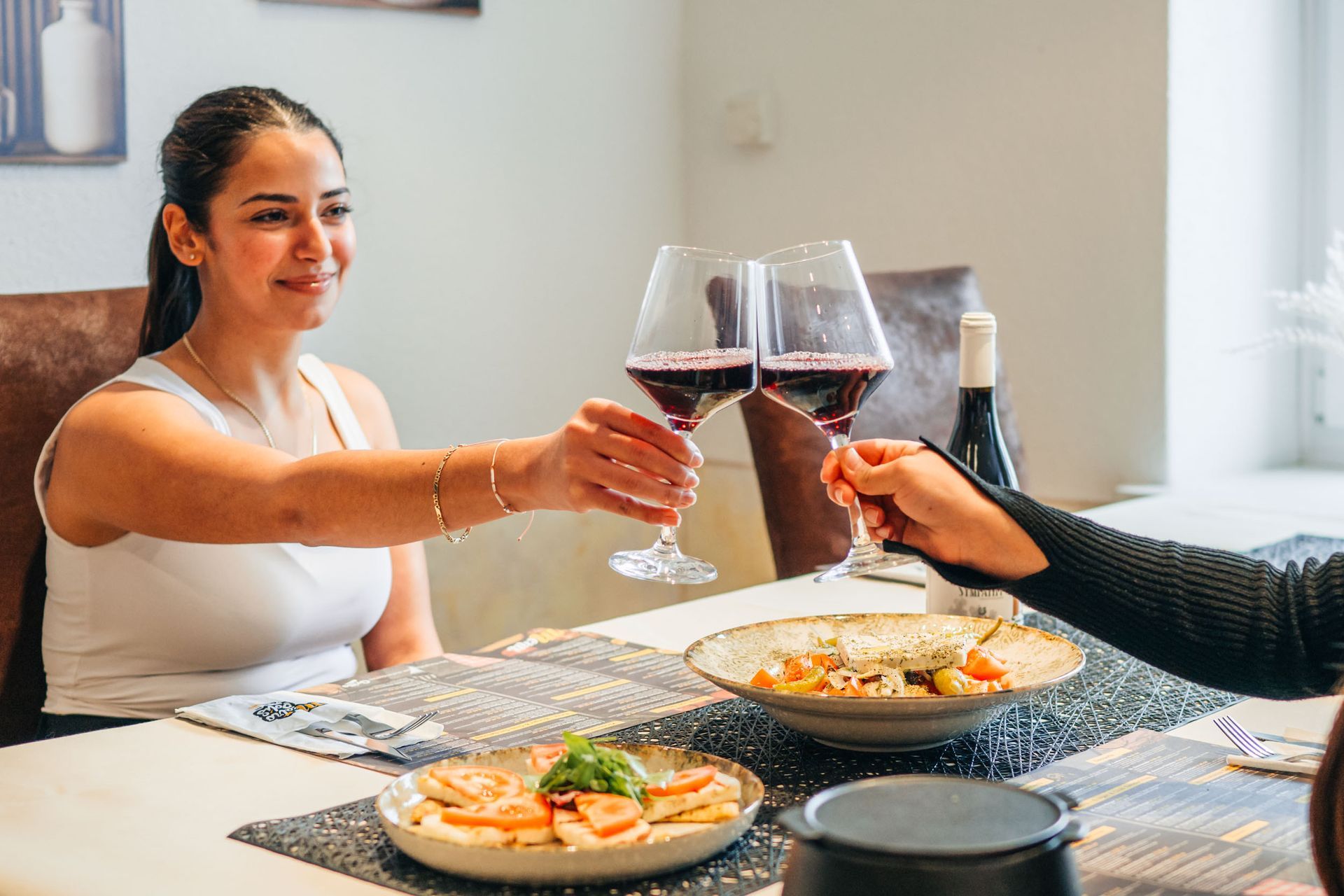 Eine Frau stösst mit einer anderen Person an einem Tisch an, beide halten Weingläser in der Hand; Speisen sind vorhanden.