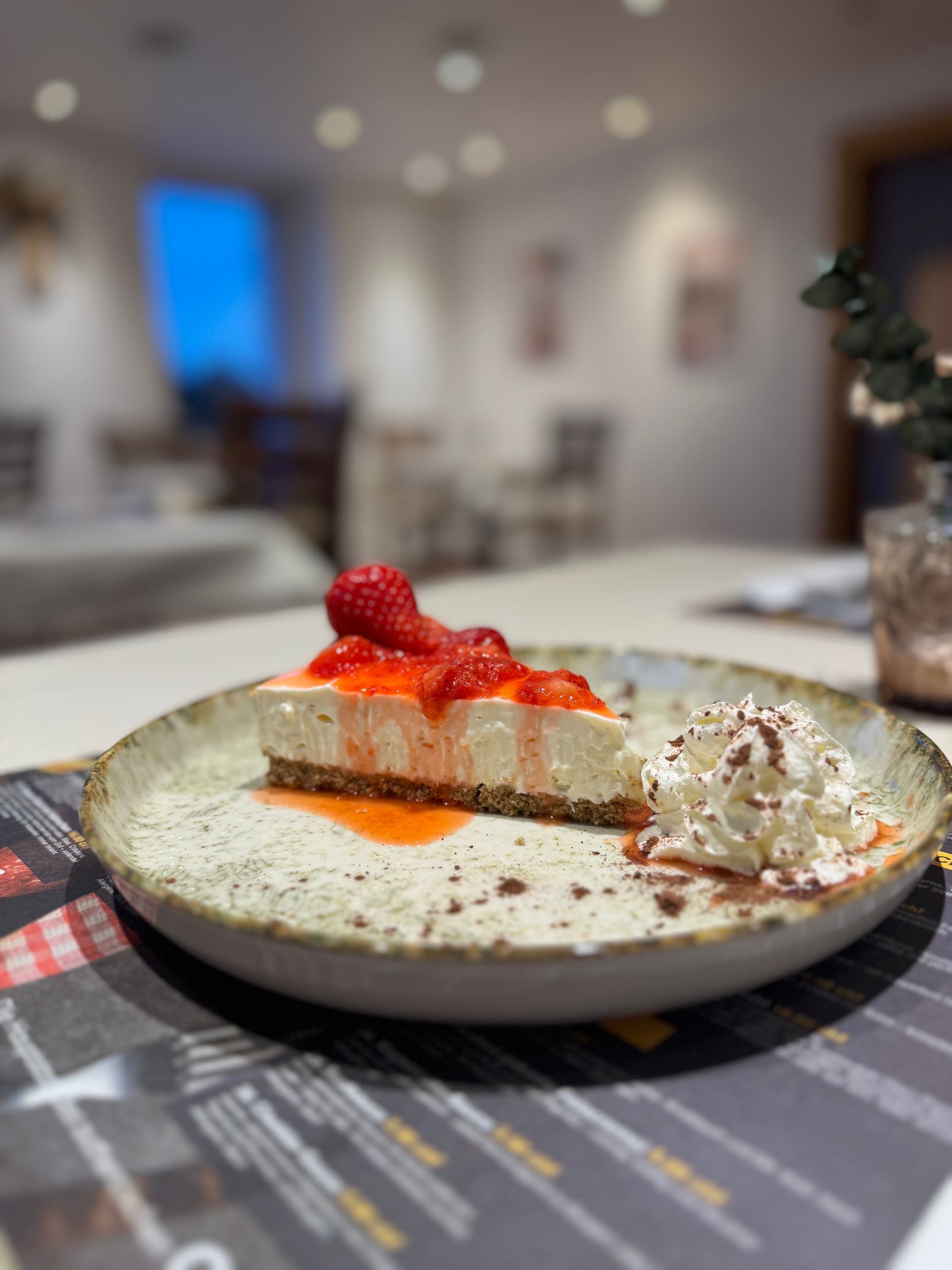Ein Stück Käsekuchen mit Erdbeersauce und Schlagsahne auf einem Keramikteller. Restaurantambiente.