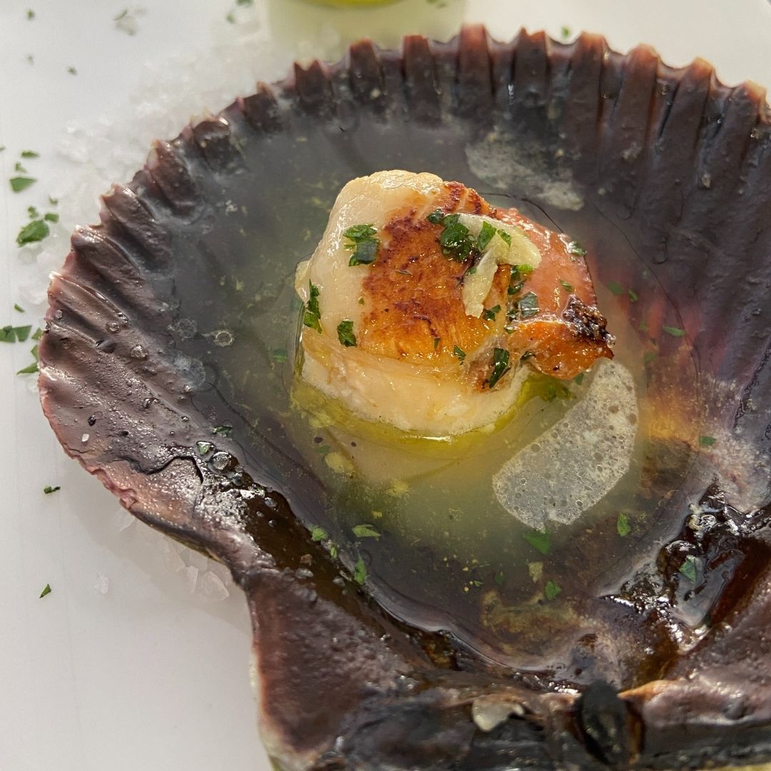 Vieira sellada en su concha con caldo y guarnición de perejil.