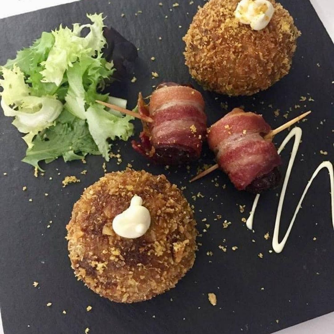 Aperitivos en pizarra negra: huevo escocés, dátiles envueltos en tocino y pasteles fritos con verduras y un chorrito de salsa.