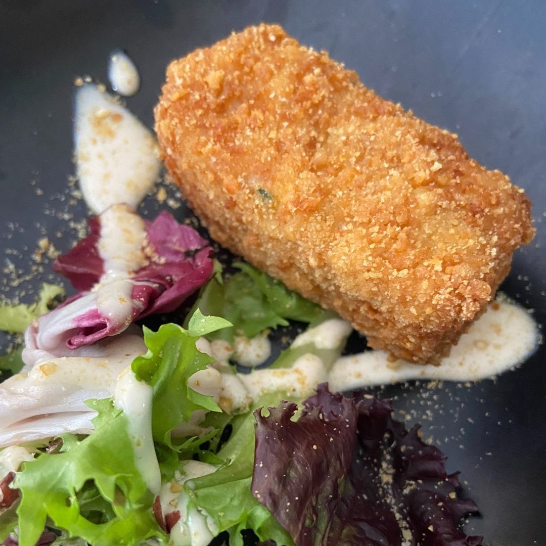 Croqueta rectangular rebozada y frita sobre una cama de lechuga, rociada con salsa, en un plato negro.