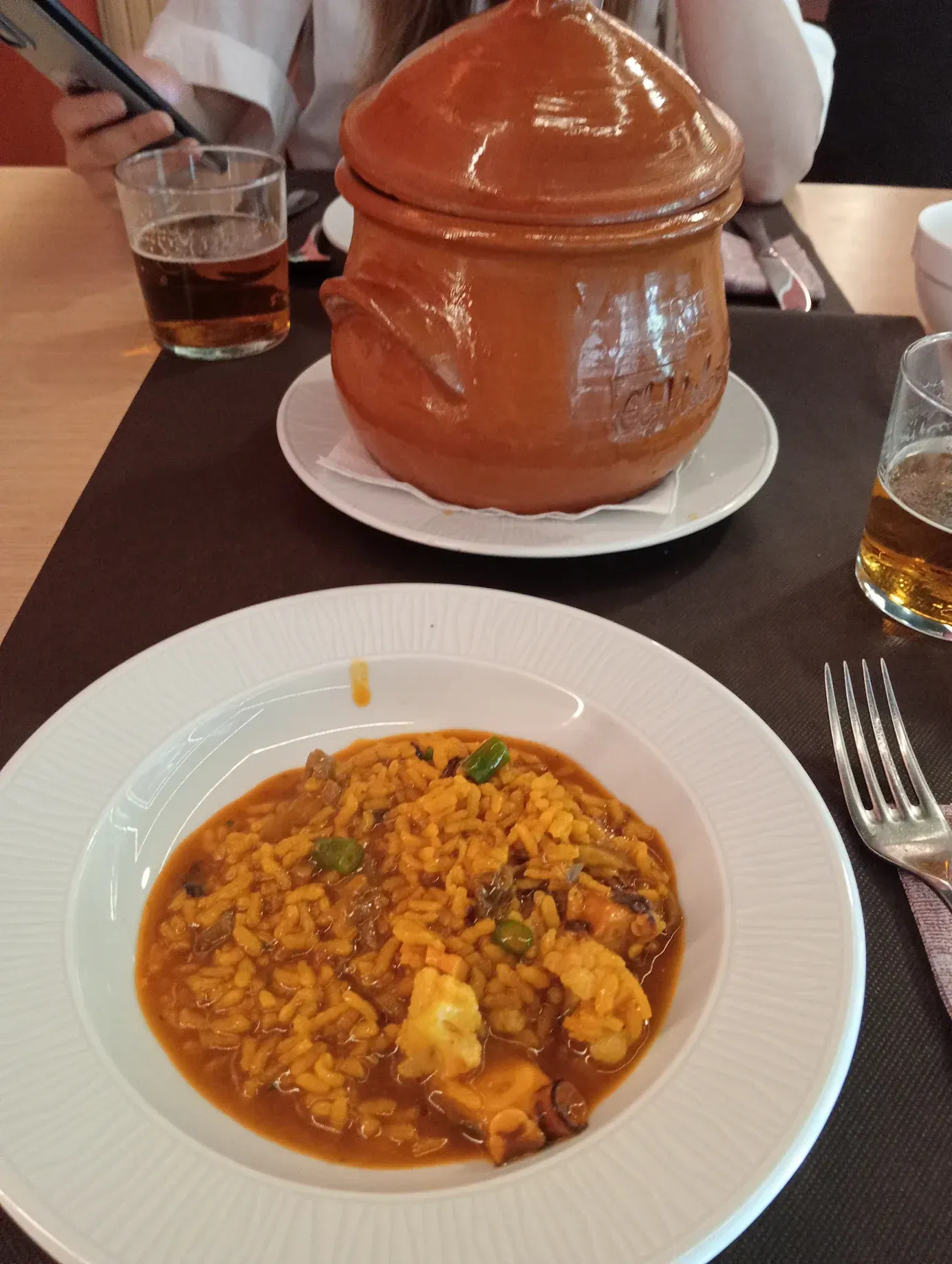 Plato de paella y una cazuela de barro con tapa sobre una mesa con bebidas y cubiertos.