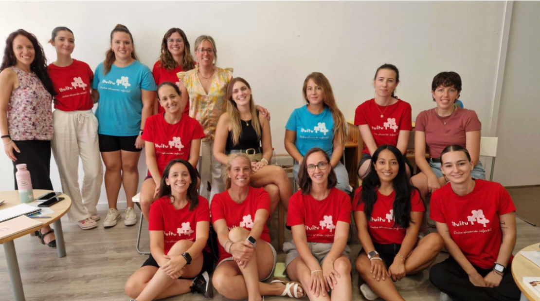 Grupo de mujeres con camisetas rojas y azules posando en una habitación. Algunas están sentadas, otras de pie.
