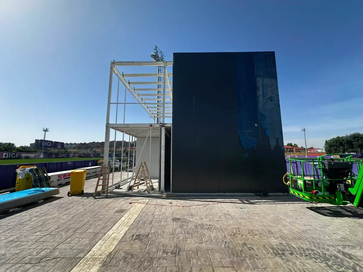 Estructura industrial al aire libre con un panel negro, marco blanco y cielo azul sobre una superficie pavimentada.