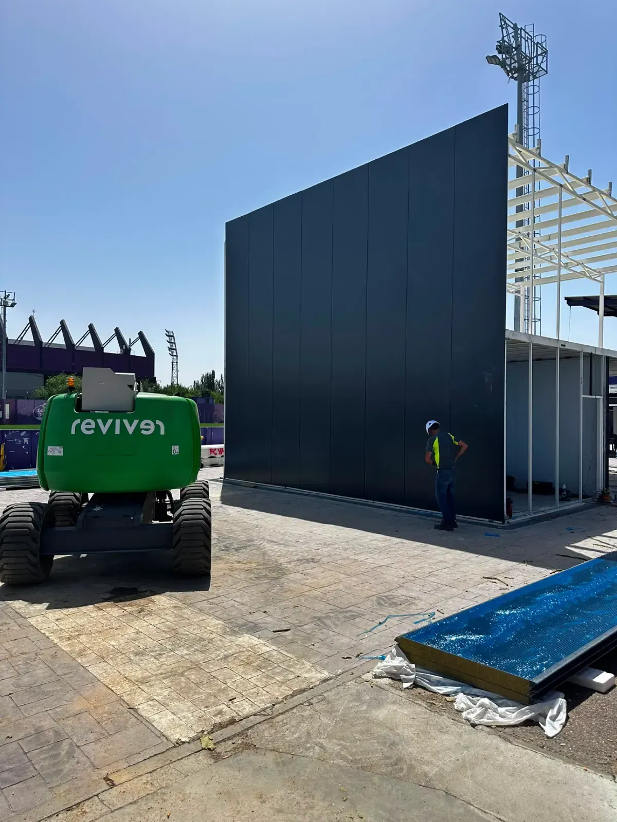 Obra en construcción con excavadora verde junto a un alto muro oscuro y una estructura de acero bajo un cielo azul claro.