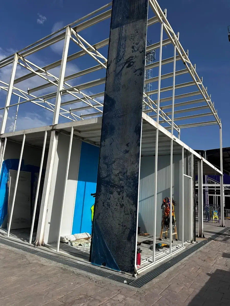 Edificio parcialmente construido con paredes blancas, puerta azul y estructura de acero a la vista bajo un cielo despejado.