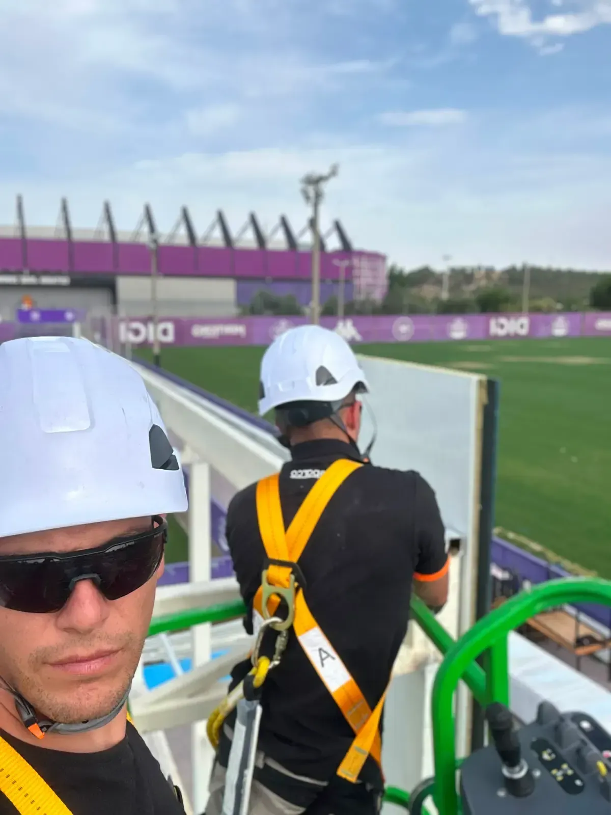 Dos trabajadores con cascos y arneses observan un estadio deportivo desde una plataforma elevada.