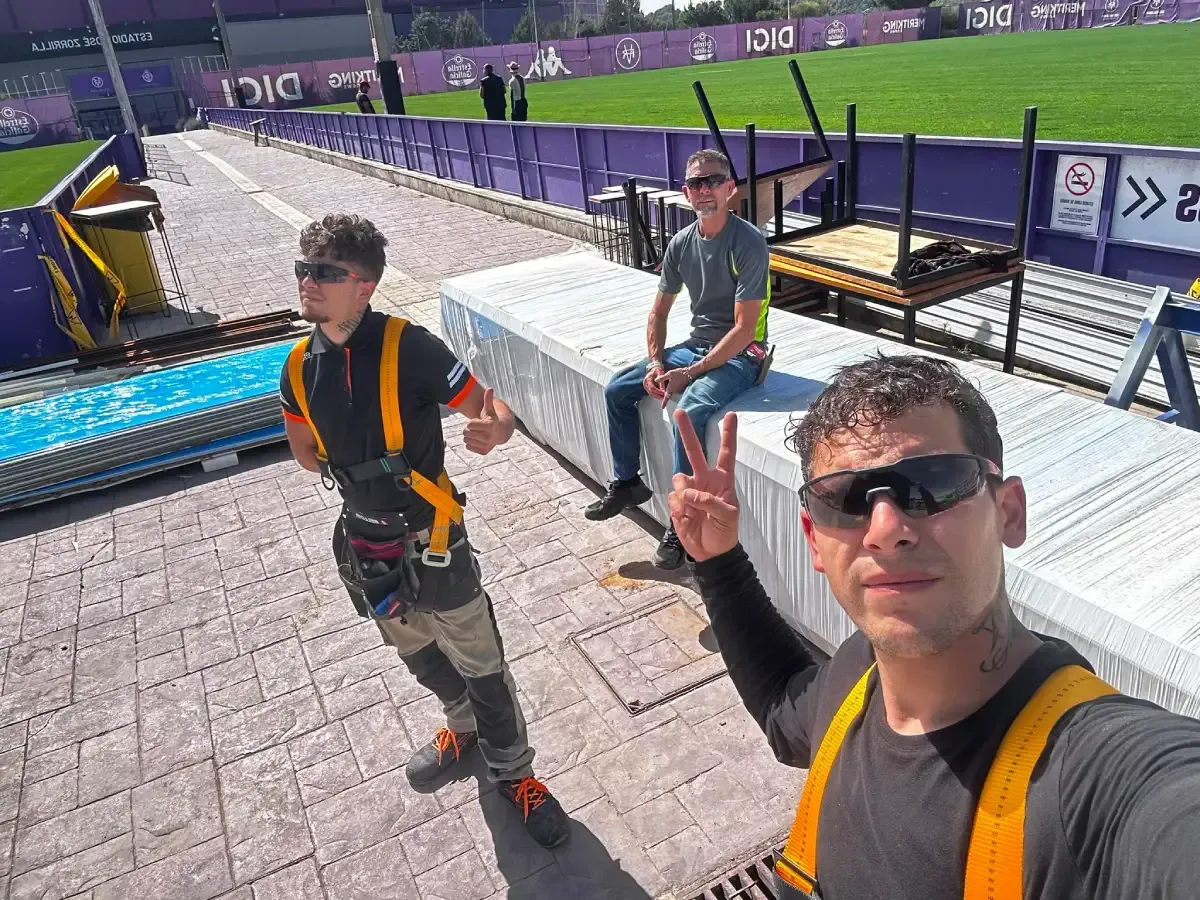 Tres personas con gafas de sol posan junto a una obra en construcción, con una grúa y césped al fondo.