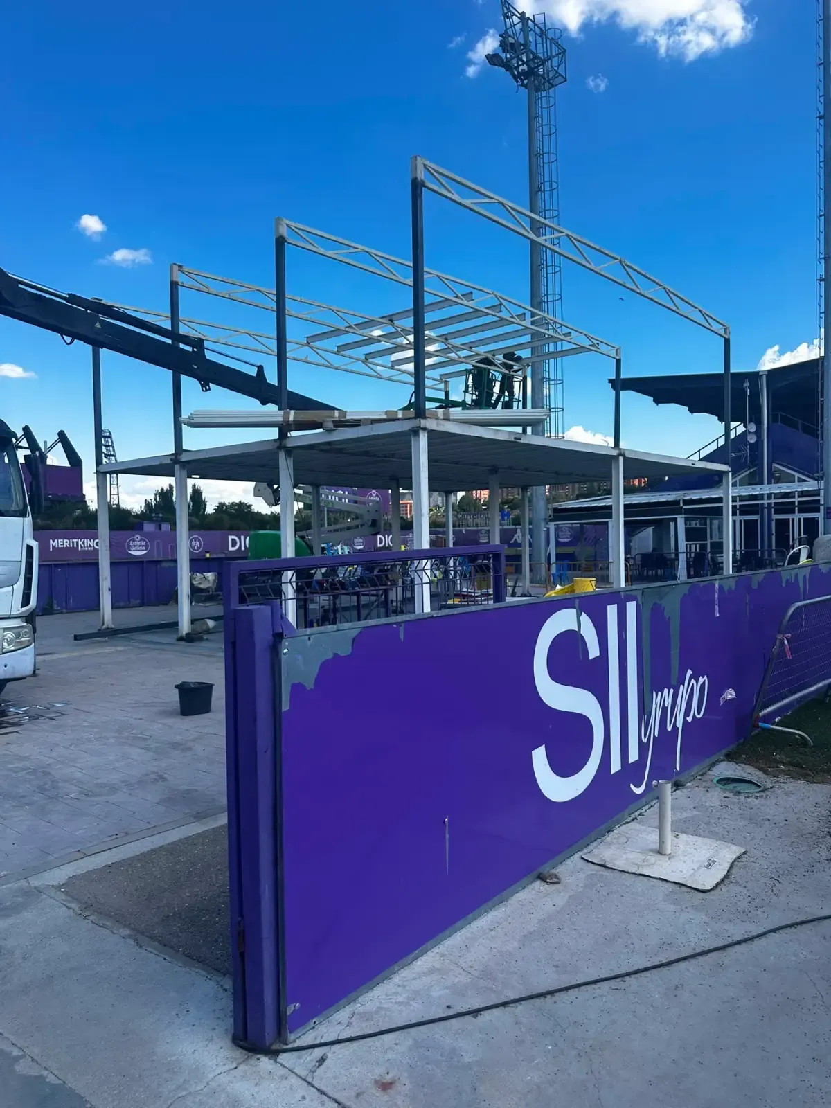 Puesto de mercado al aire libre con barreras moradas y estructura metálica bajo un cielo azul brillante.
