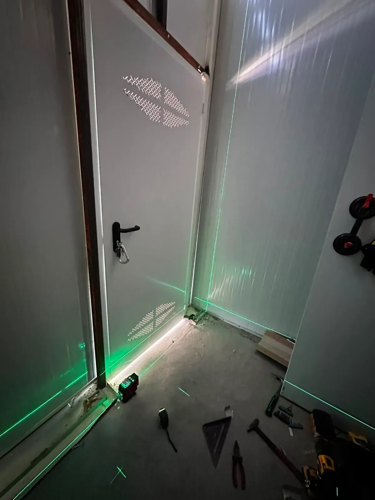 Puerta blanca entreabierta con luces verdes en el suelo en una habitación estrecha; herramientas esparcidas por el suelo.