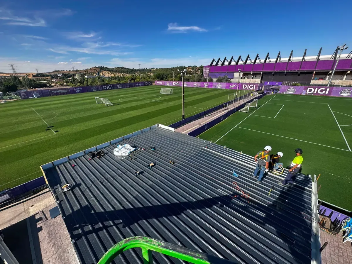 Estadio de fútbol con césped verde, gradas moradas y vistas desde la azotea bajo un cielo despejado.