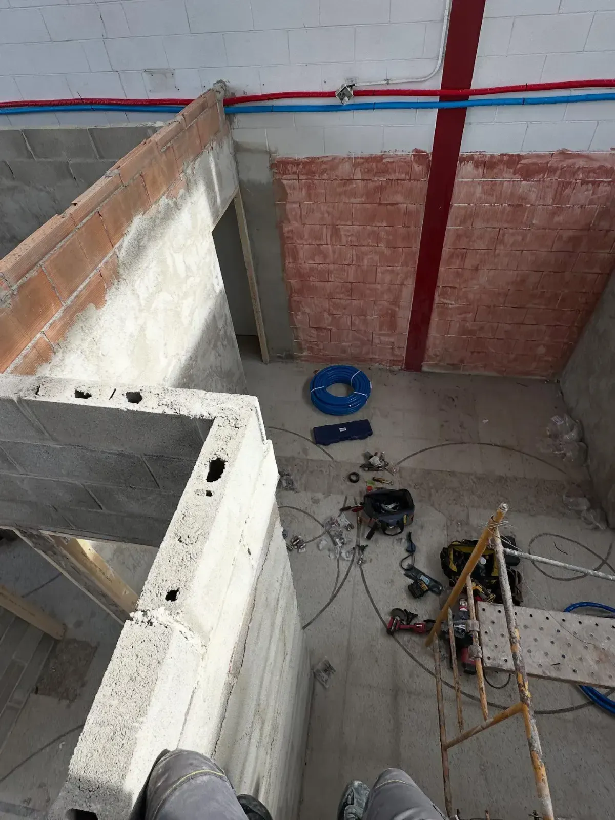 Zona de construcción interior con paredes sin terminar, herramientas, cables y un cubo azul en el suelo.