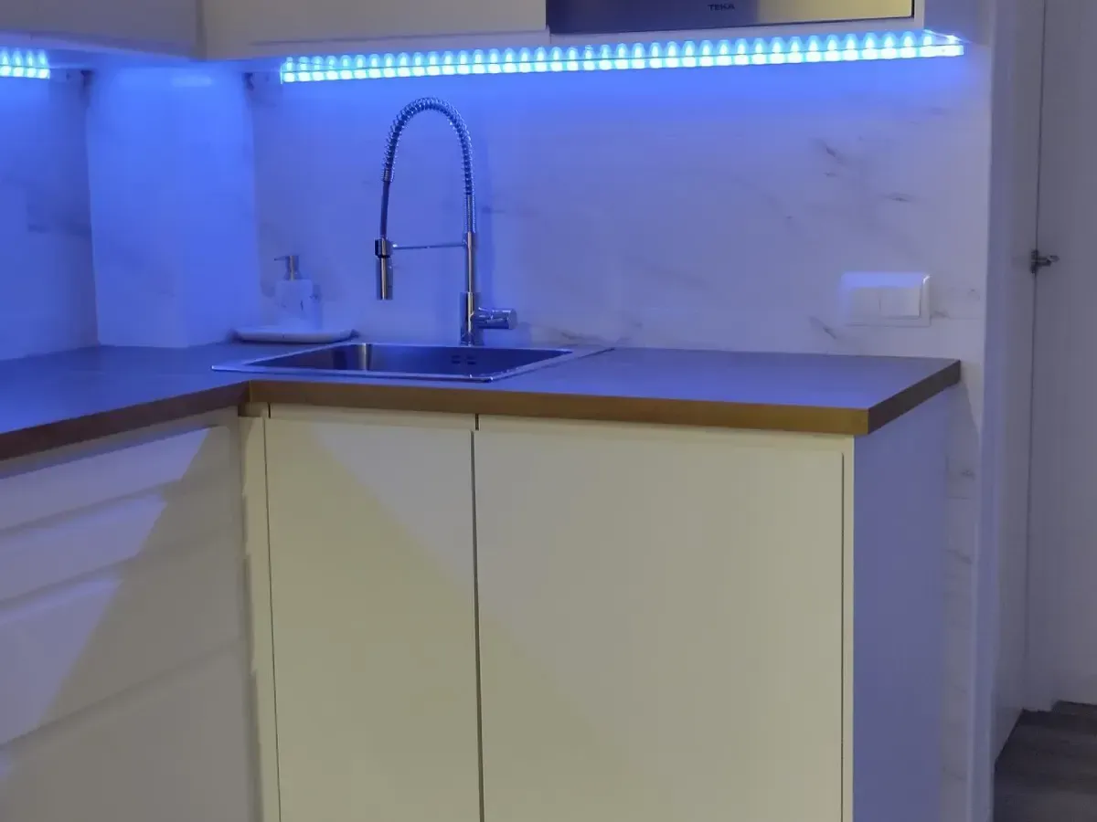 Pequeño rincón de cocina con fregadero, encimera de madera, armarios color crema e iluminación LED azul bajo los armarios.