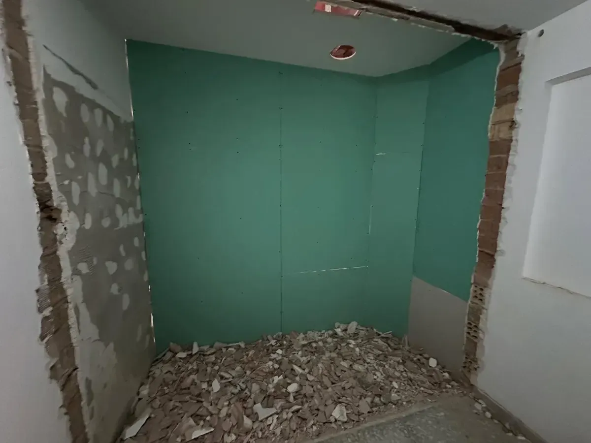 Habitación parcialmente reformada con paredes de pladur verde, yeso visto y un montón de escombros en el suelo.