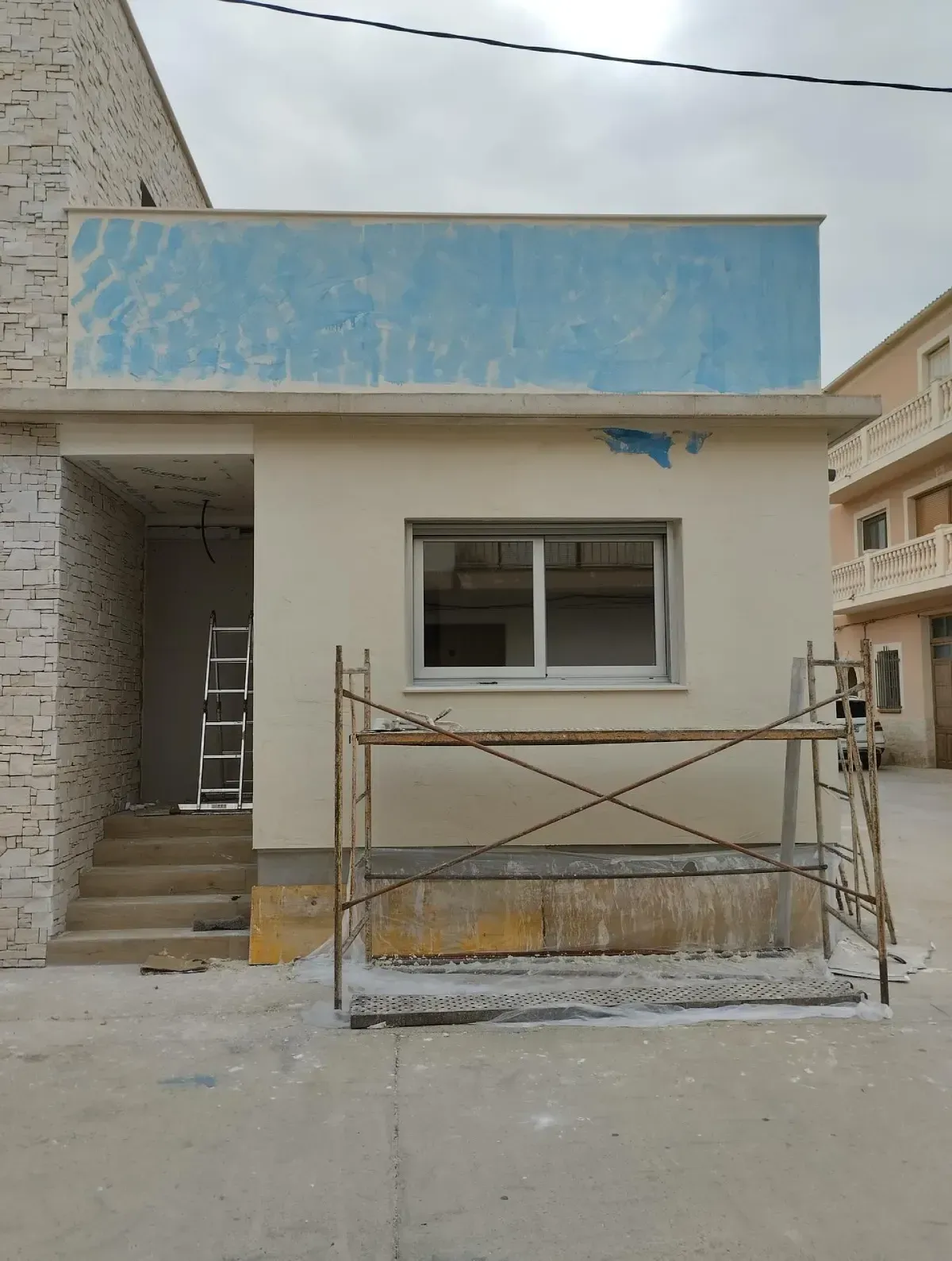 Exterior de una pequeña casa sin terminar con fachada azul, escaleras delanteras, ventana y andamios en la parte frontal.