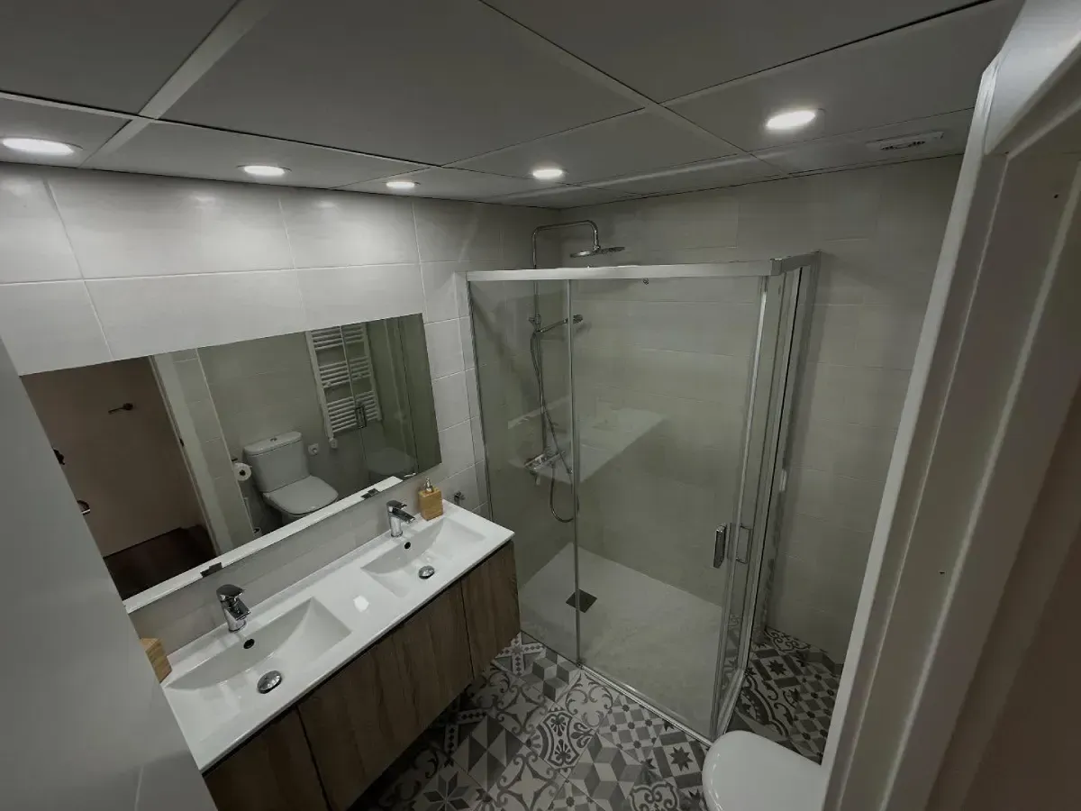 Baño moderno con lavabo doble, espejo grande, ducha de cristal y suelo de baldosas estampadas.