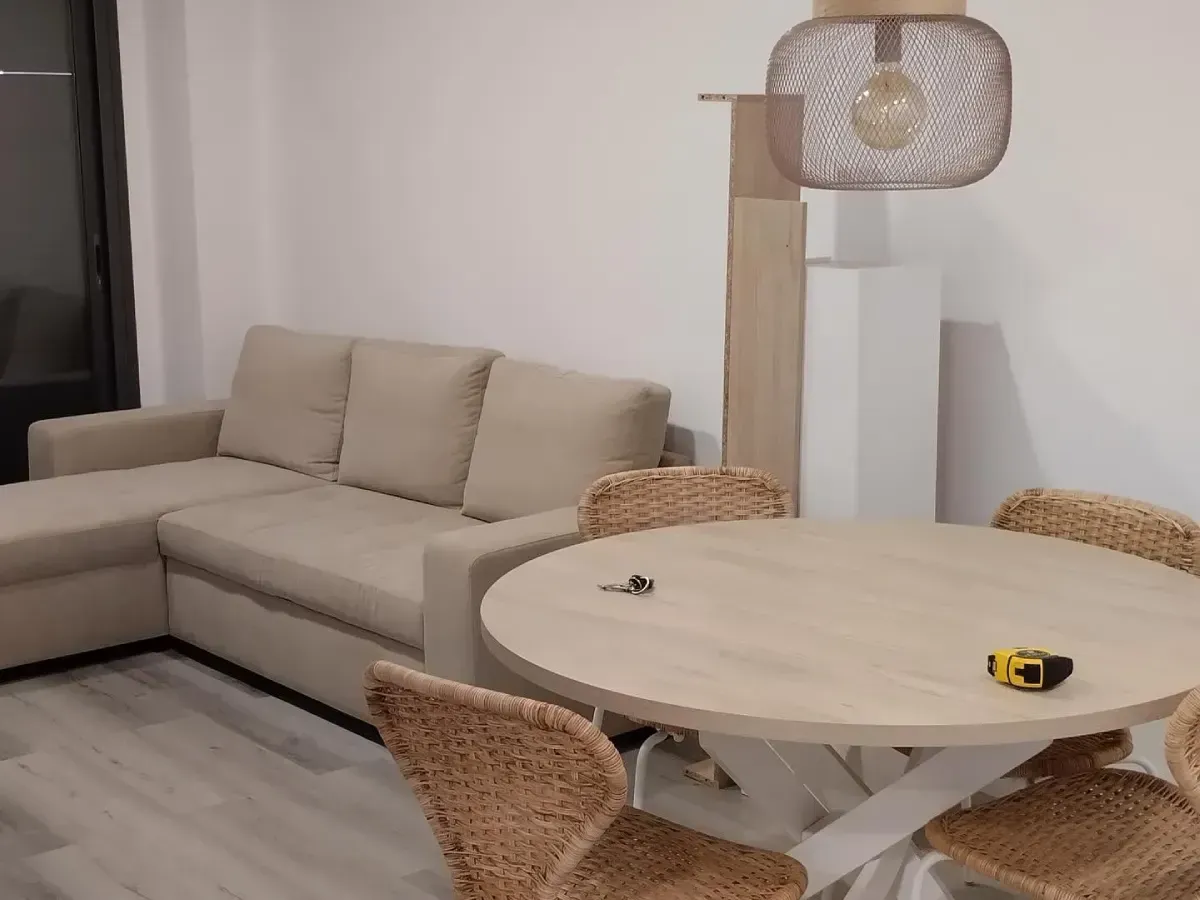 Sala de estar moderna con sofá seccional beige, mesa de comedor redonda, sillas de mimbre y lámpara de pie.