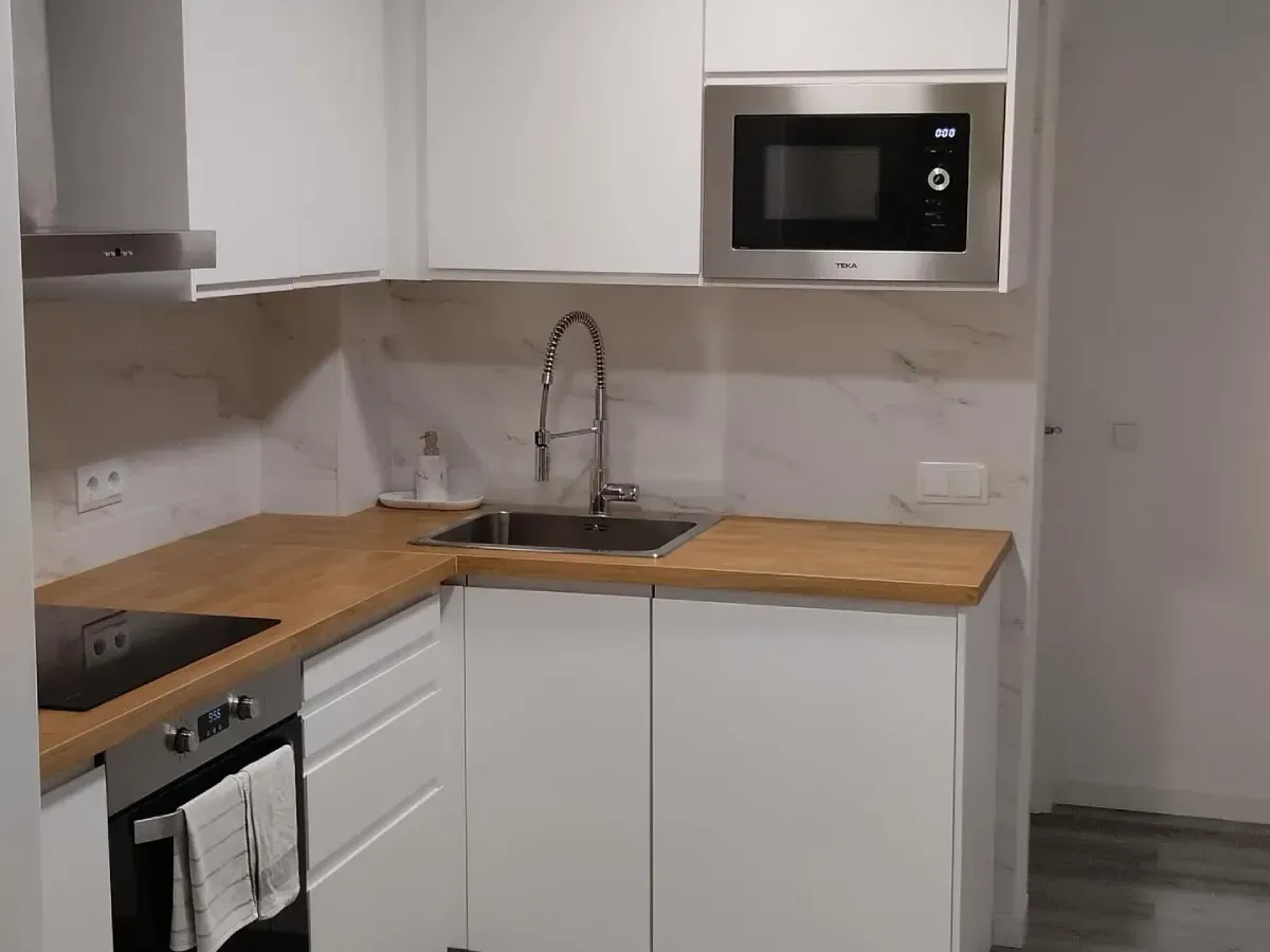 Pequeña cocina moderna con armarios blancos, encimeras de madera, fregadero, fogones y microondas.