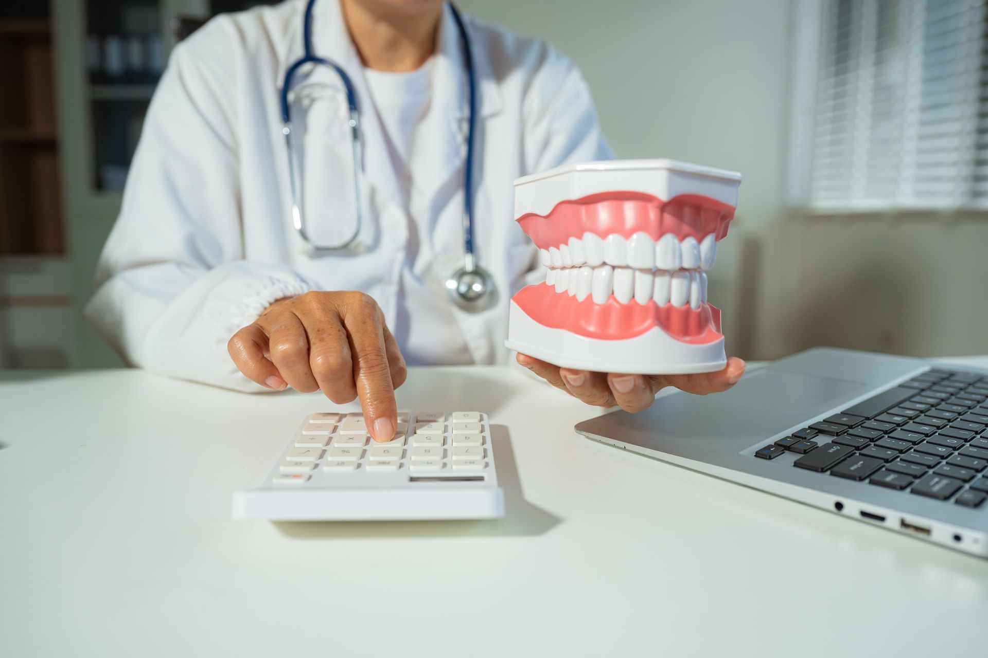 Ein Mediziner im Laborkittel benutzt einen Taschenrechner, während er neben einem Laptop ein Modell menschlicher Zähne hält.