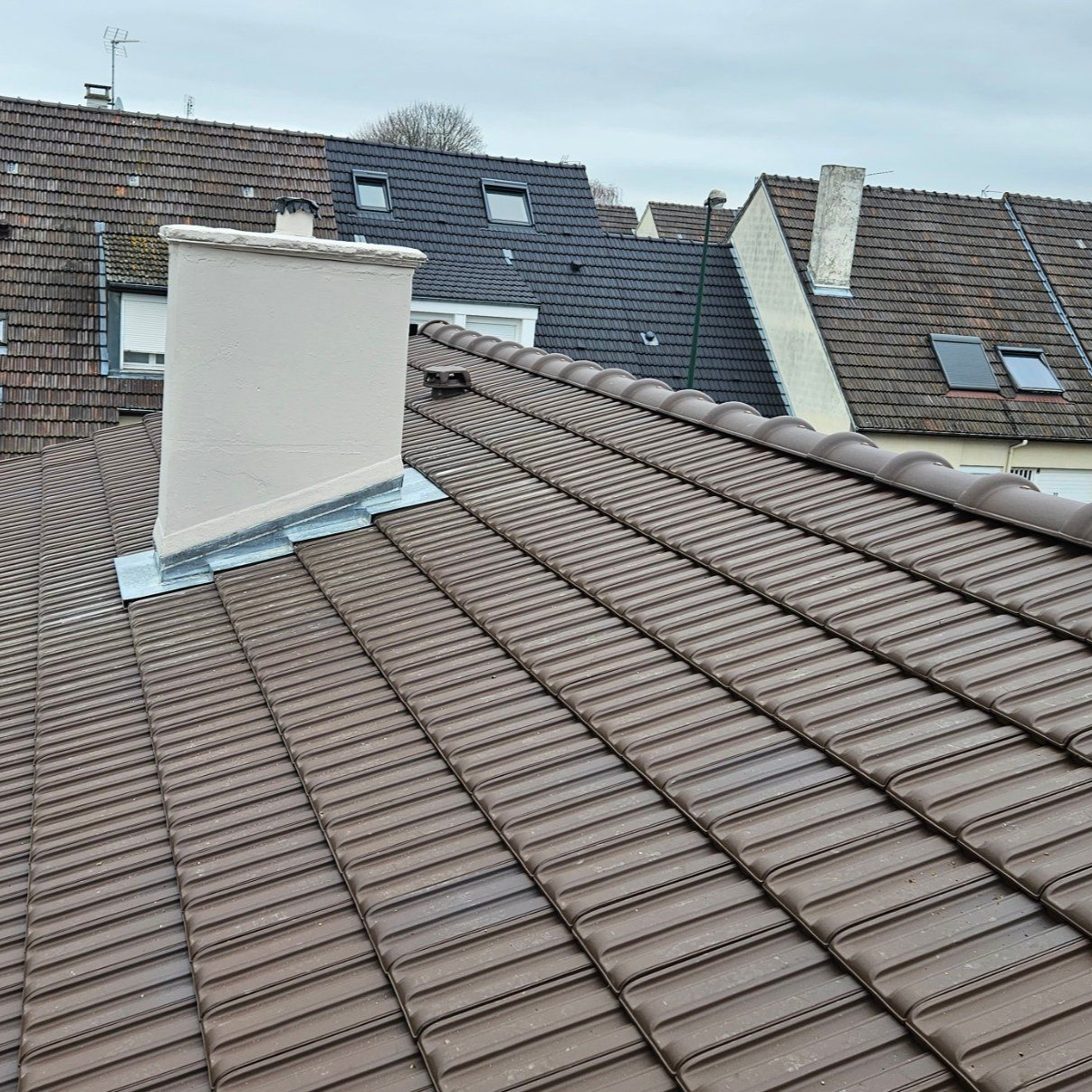 Toits de tuiles brunes avec une cheminée beige, ciel gris couvert.