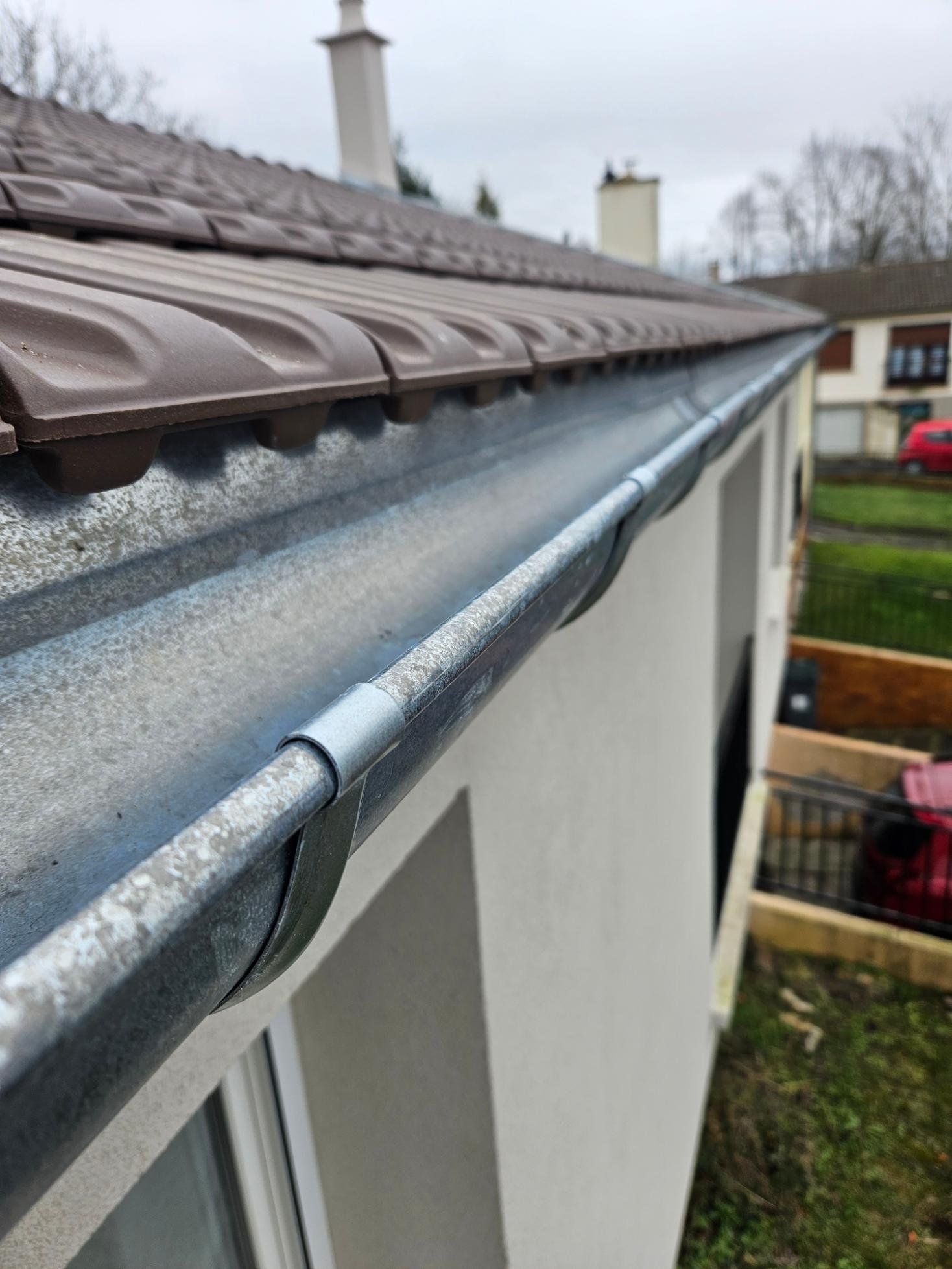 Gros plan sur le toit d'un bâtiment avec des tuiles brunes, une gouttière argentée et un mur blanc.