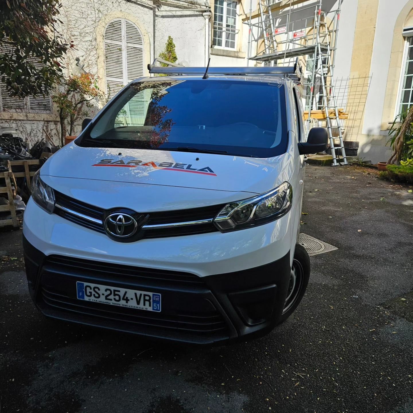 Une camionnette Toyota blanche portant le logo de l'entreprise est garée devant un bâtiment ; des échafaudages sont visibles.