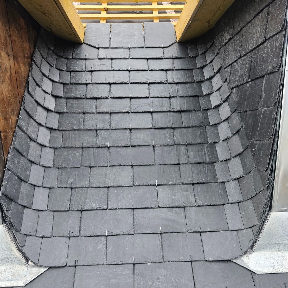 Gros plan sur une section de toit en ardoise incurvée, les bardeaux gris foncé étant disposés en rangées sous des poutres en bois.