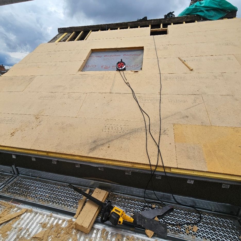 Chantier de construction : une tronçonneuse sur la façade d’un bâtiment avec une fenêtre ouverte et une structure en bois apparente.