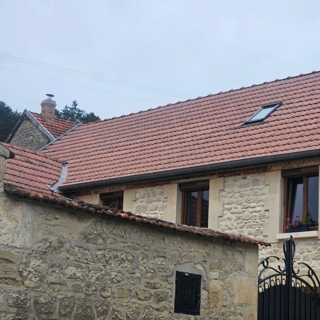 Maison en pierre avec toit de tuiles rouges, petite fenêtre et volets sombres.