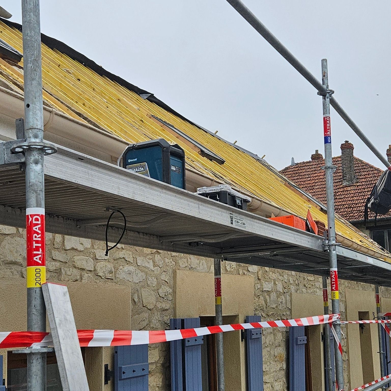 Échafaudage à côté d'un bâtiment au toit jaune partiellement exposé, ruban de signalisation rouge et blanc.