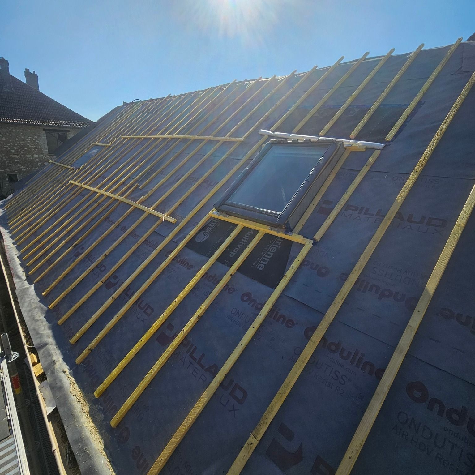 Toiture en construction avec puits de lumière, membrane noire et lattes de bois, sur fond de ciel bleu.