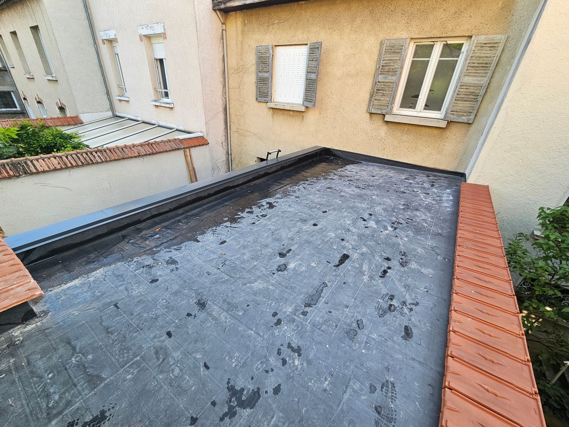 Toit plat et sombre bordé de briques. Bâtiments et fenêtres en arrière-plan. Journée ensoleillée.