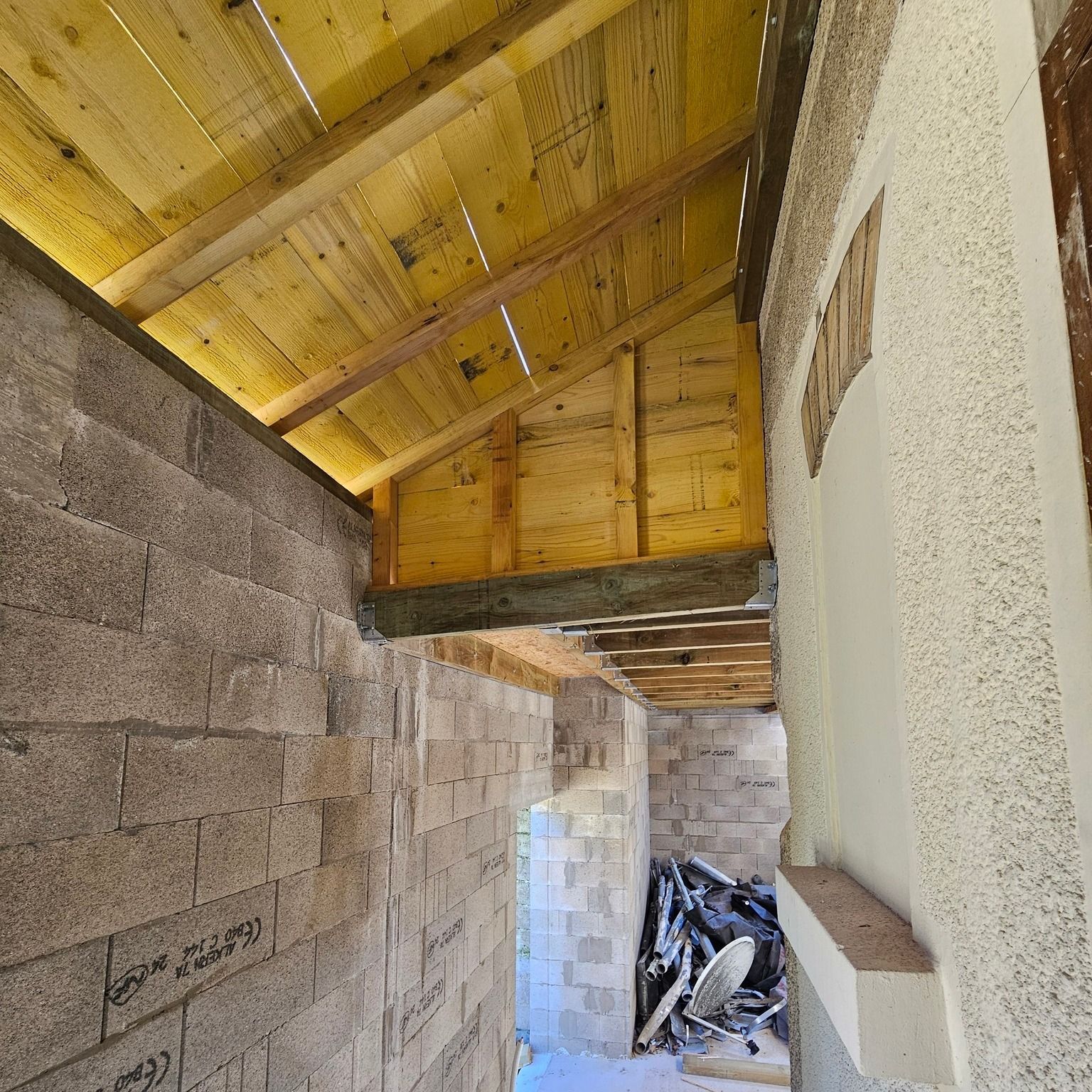 Construction de l'intérieur d'un bâtiment inachevé. Le plafond en bois apparent et les murs en parpaings sont visibles.