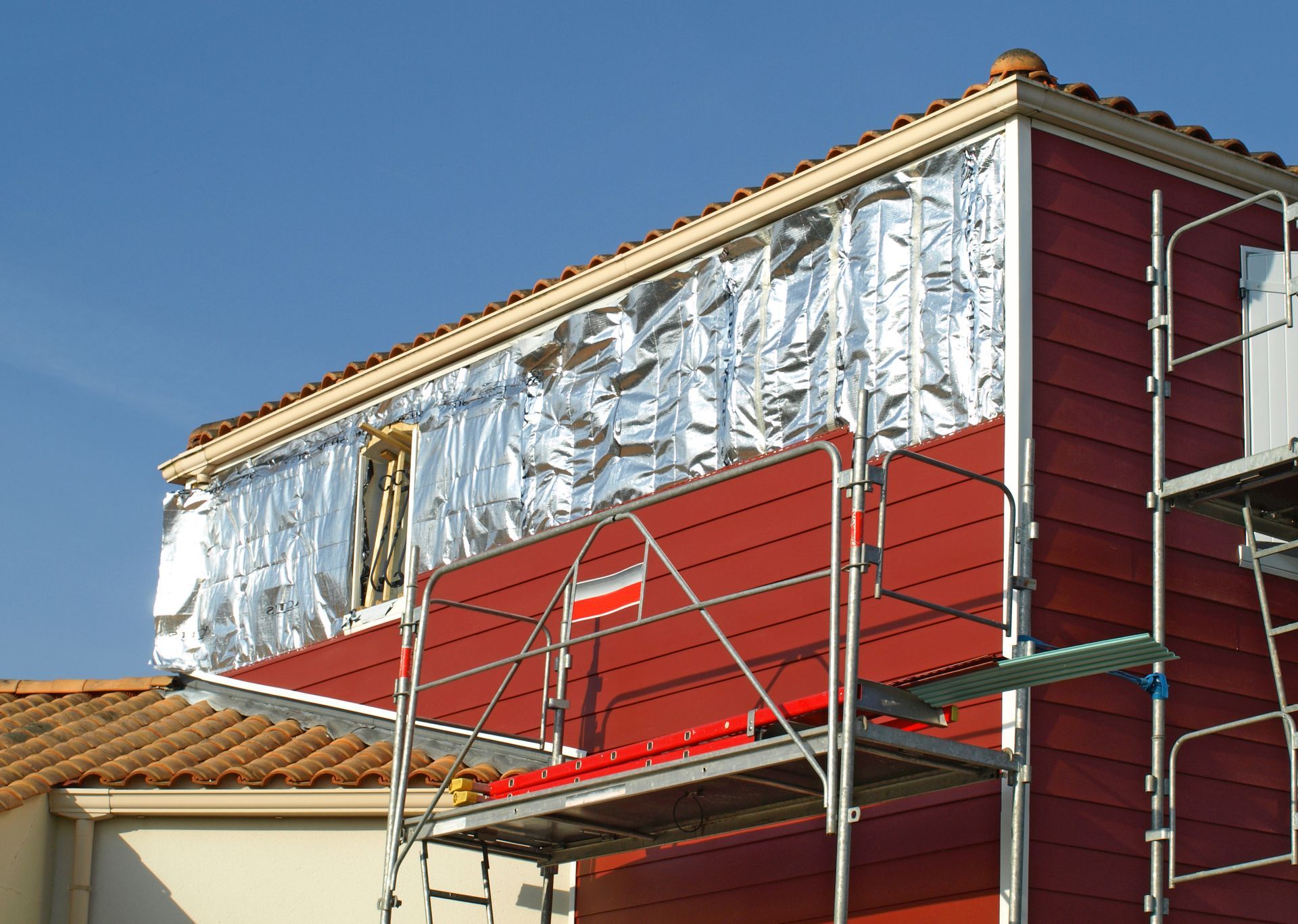 Chantier de construction avec bardage rouge, isolation et échafaudages sur fond de ciel bleu.