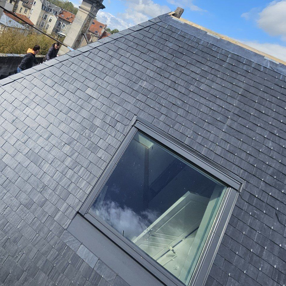 Un toit en ardoise percé d'un puits de lumière, ciel bleu. Deux personnes travaillent sur le toit.
