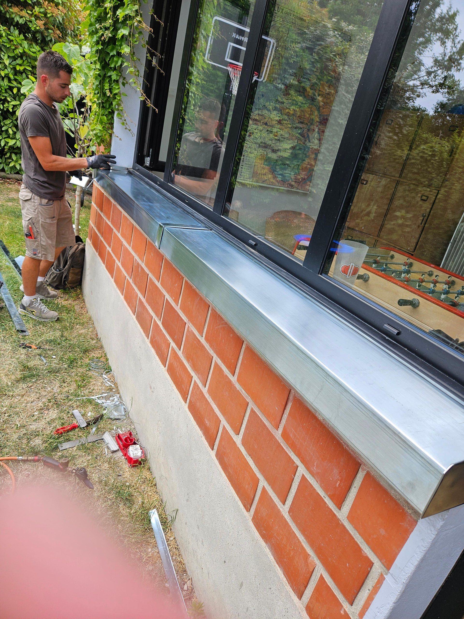 Un homme installe des solins métalliques au-dessus d'un mur de briques et d'une fenêtre, à l'extérieur.