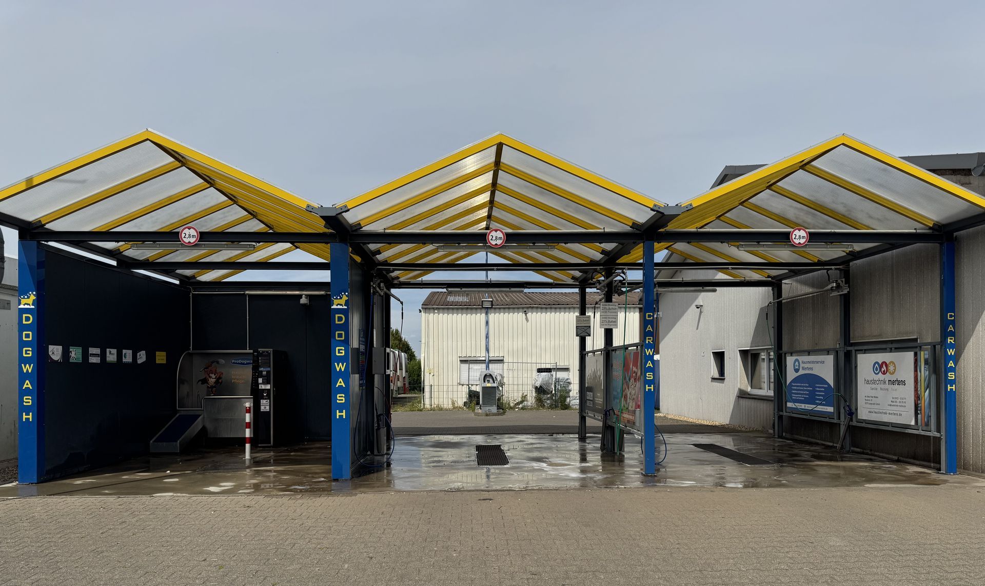 Autowaschanlage mit blau-gelbem Vordach, nasser Asphalt.
