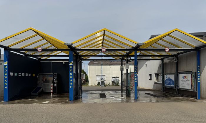 Autowaschanlage mit blau-gelbem Vordach, nasser Asphalt.