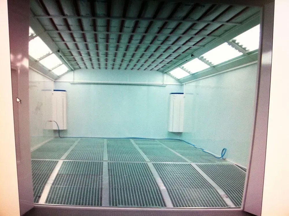 Eine leere, gut beleuchtete Spritzkabine. Zu sehen sind Gitterboden, hellblaue Wände und eine Rasterdecke.