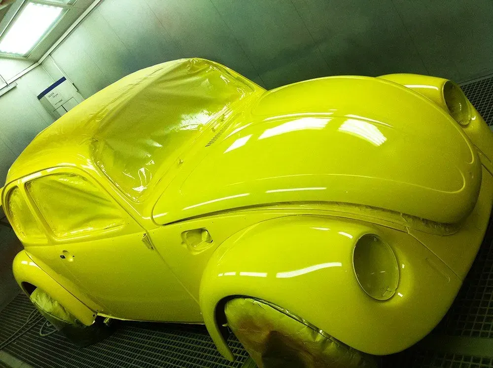 Gelber Volkswagen Käfer wird in einer Spritzkabine neu lackiert. Das Auto ist mit frischer Farbe bedeckt.