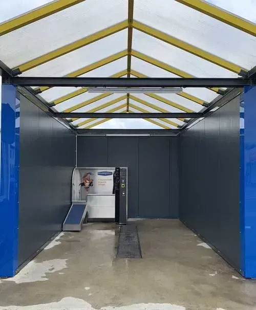 Innenansicht einer Autowaschanlage mit einer silbernen Maschine, grauen Wänden und einem gelben und durchsichtigen Dach. Blaue Säulen rahmen den Eingang ein.