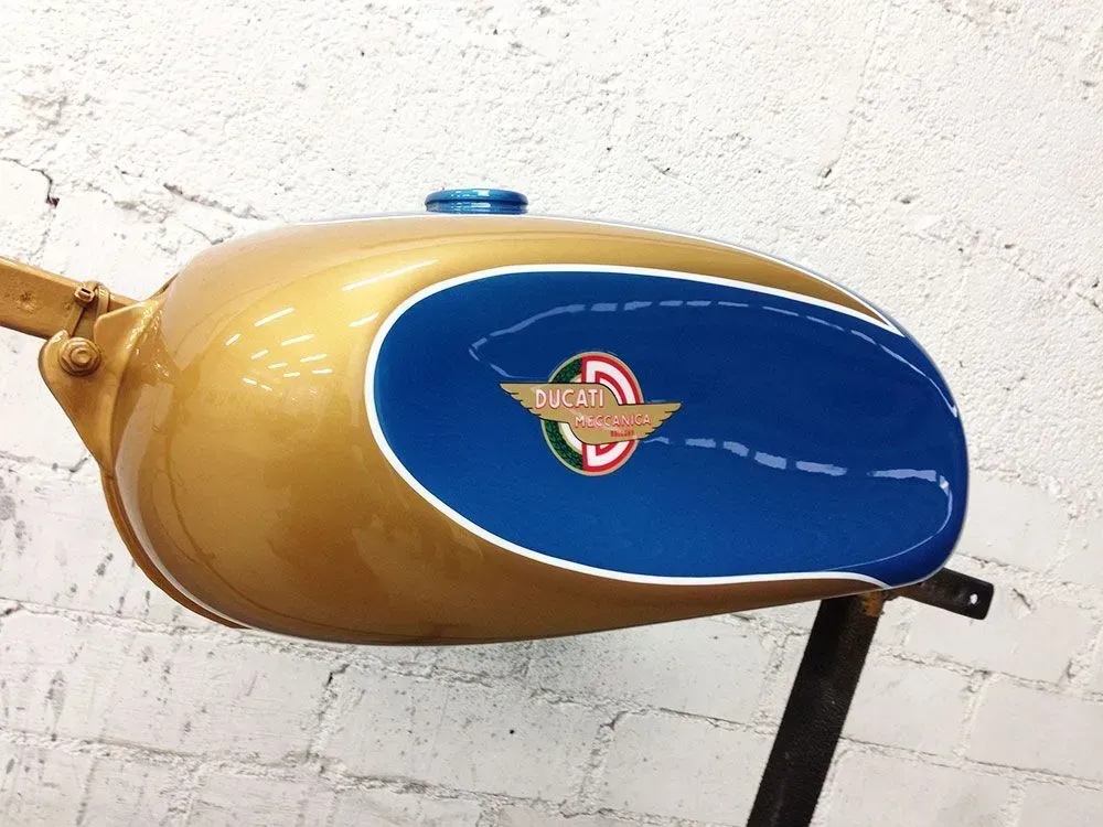 Gold- und blauer Vintage-Motorrad-Benzintank mit Logo und Tankdeckel, an einer weißen Ziegelwand montiert.