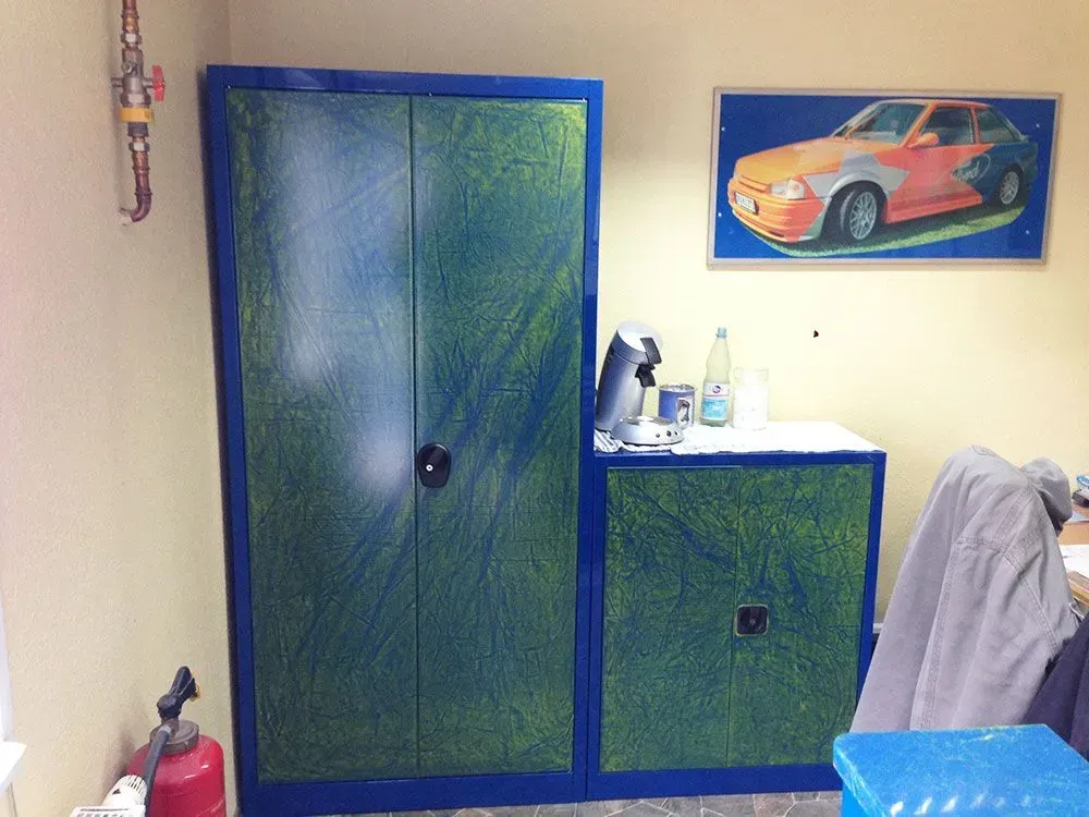Ein grün und blau gestrichener Schrank und eine kleine Truhe stehen neben einer weißen Fläche mit Flaschen und einer Kaffeemaschine. An der Wand hängt eine Autozeichnung.