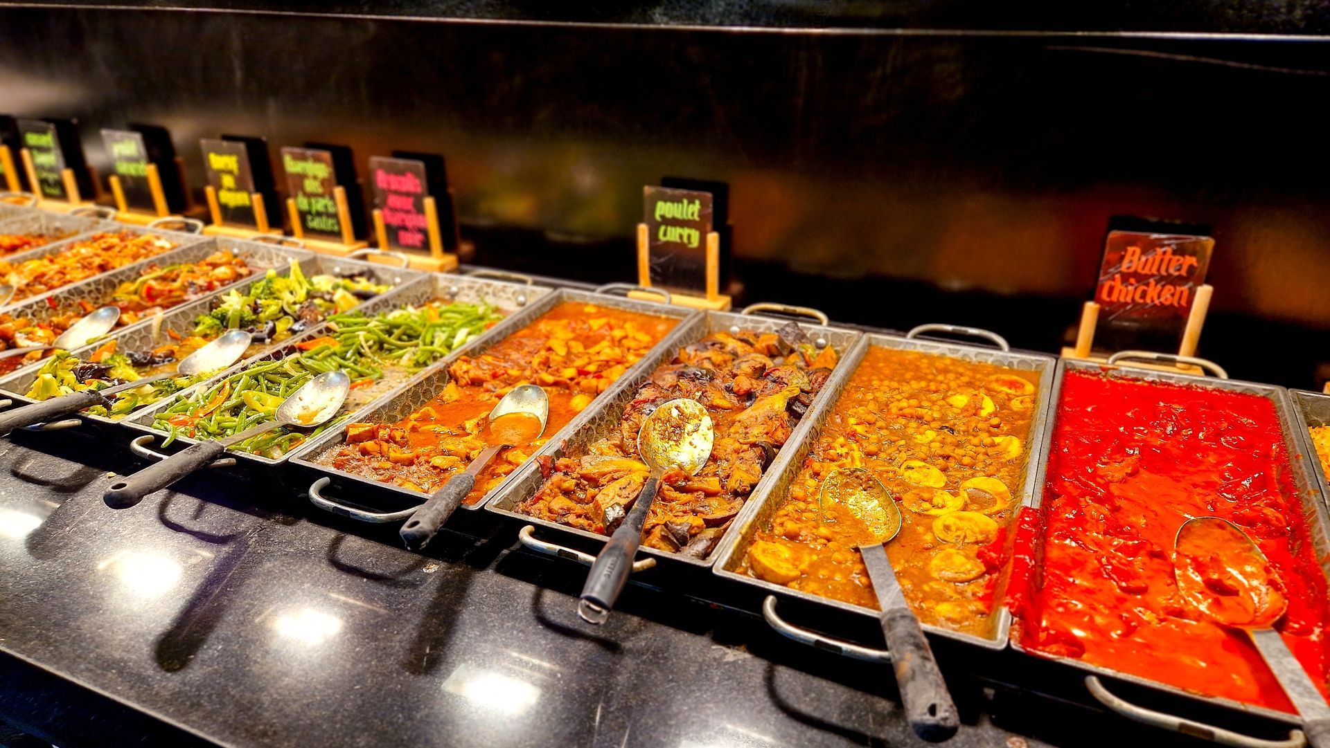 Buffet composé de divers plats indiens présentés dans des plateaux en acier inoxydable étiquetés.
