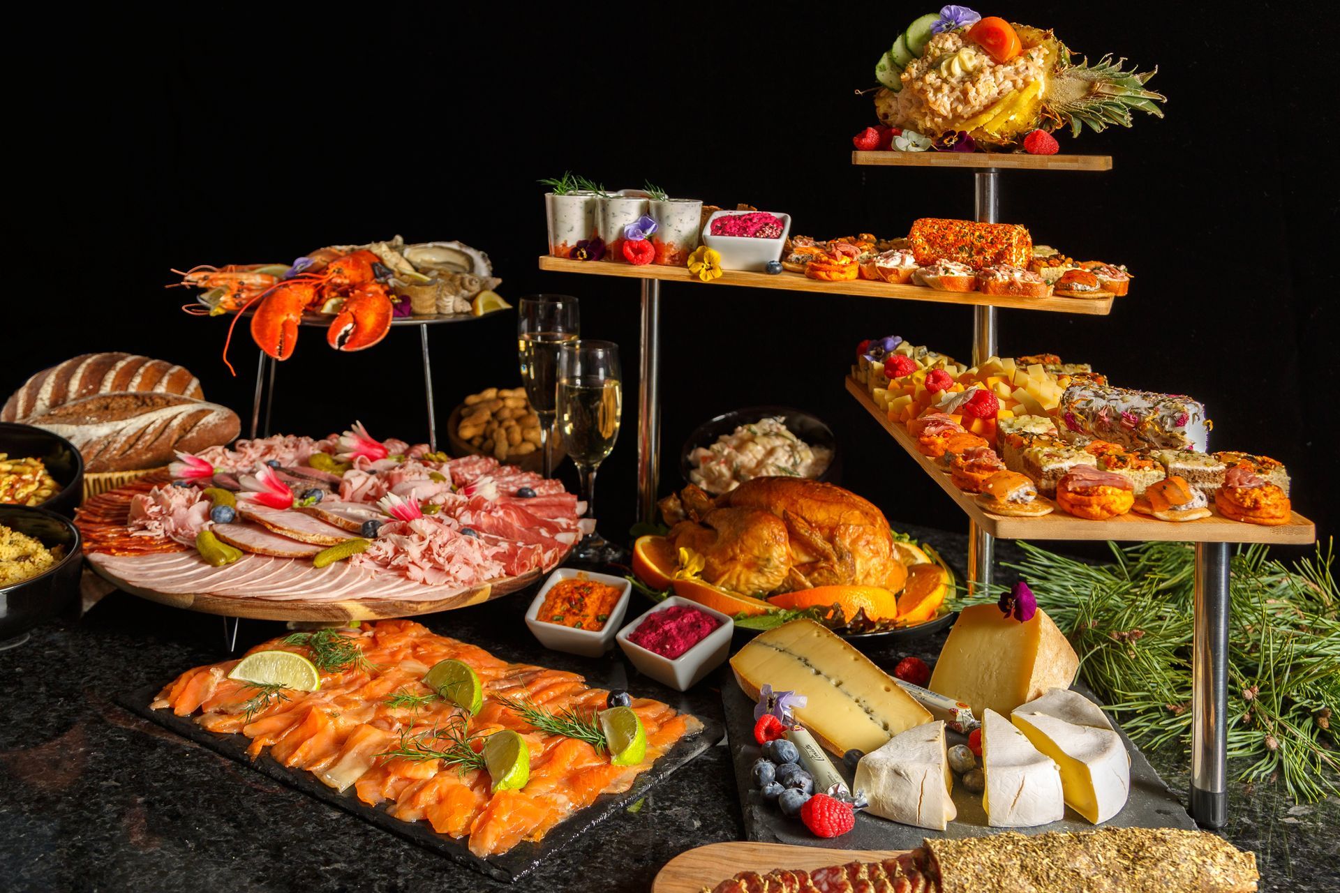Un somptueux buffet, dressé sur une table noire, propose fruits de mer, viandes, fromages, amuse-gueules et pain, le tout éclairé sur fond noir.