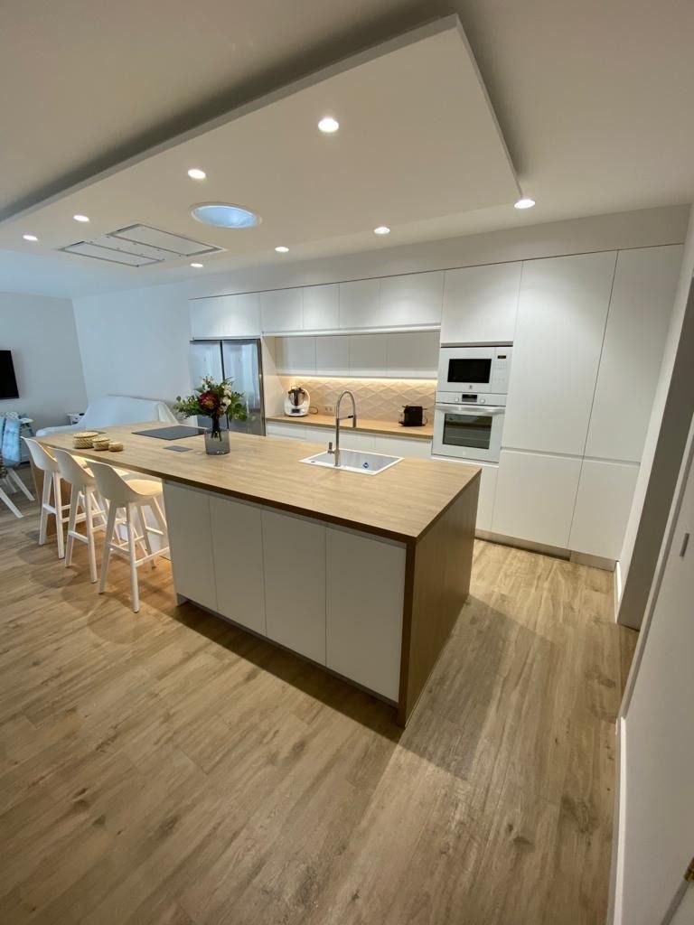 Cocina moderna blanca con suelos de madera, isla grande, taburetes de bar y electrodomésticos integrados.