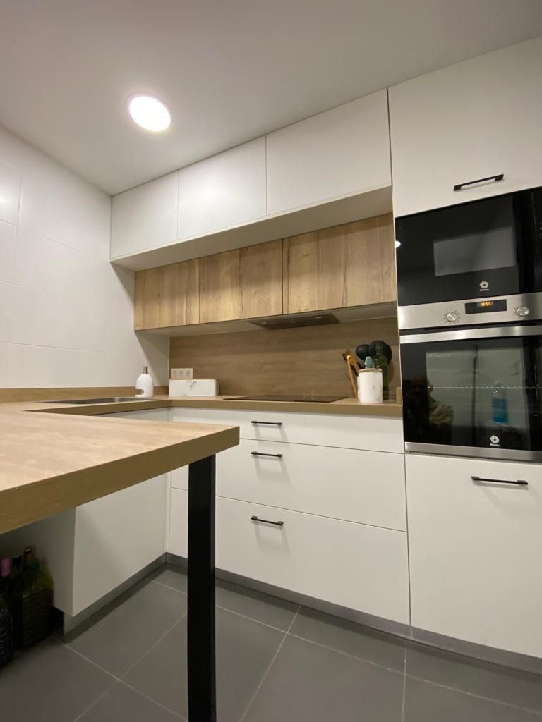 Cocina moderna blanca con detalles de madera, horno empotrado negro y una mesa de madera en primer plano.