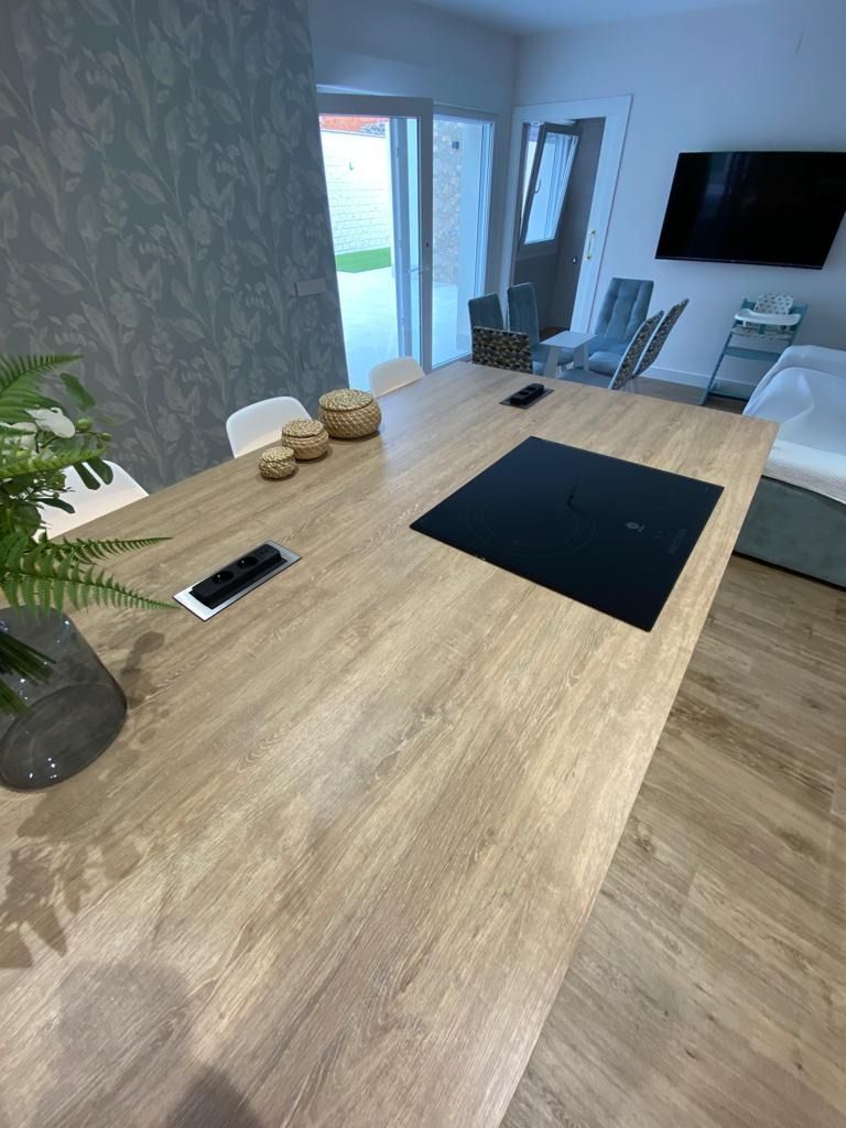 Cocina moderna blanca con detalles de madera, horno empotrado, suelo de baldosas grises y una mesa de madera en primer plano.