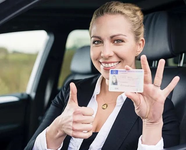 Una mujer está sentada en un automóvil sosteniendo una licencia de conducir y haciendo un gesto de aprobación con el pulgar hacia arriba.