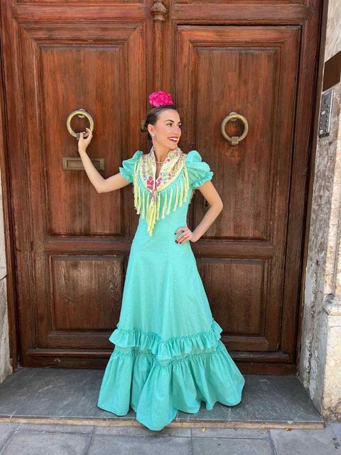 Mujer con vestido flamenco turquesa posando frente a una puerta de madera tallada, sosteniendo una anilla como tirador.